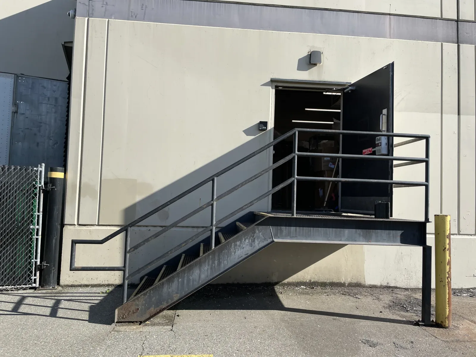 Black-painted exterior platform stair at a Vancouver loading bay with pipe-rail guards, bar grating treads, and a building-mounted landing, a current code-compliant replacement of an older pan-tread stair