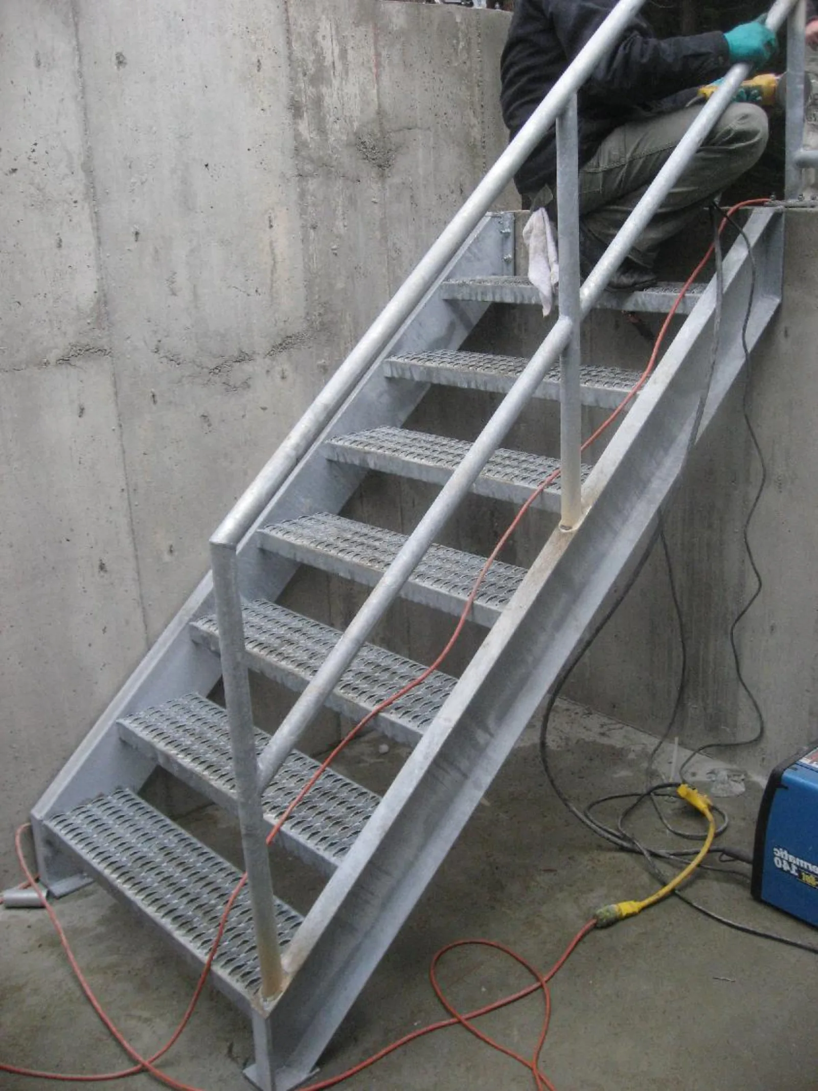 Galvanized exterior steel access stair under construction with dimple-pattern serrated plate treads and pipe-rail guards mid-install in a concrete basement well
