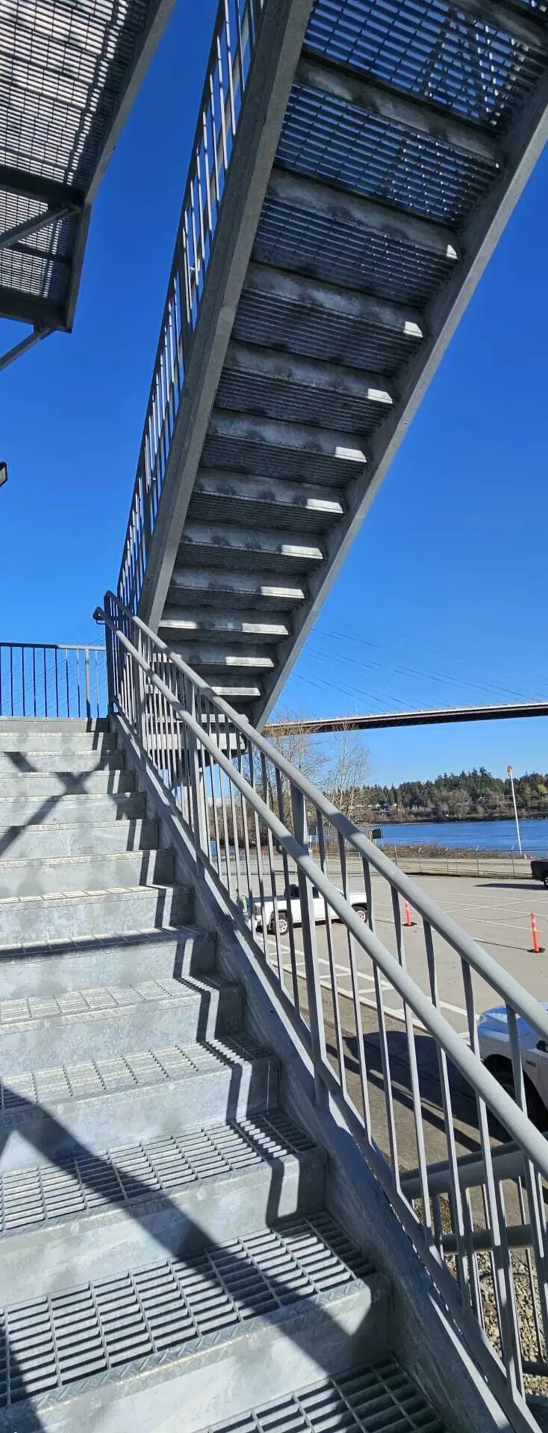 Galvanized switchback warehouse access stair against a waterfront industrial building with serrated bar grating treads and pipe-rail guards, photographed from below against a clear blue sky