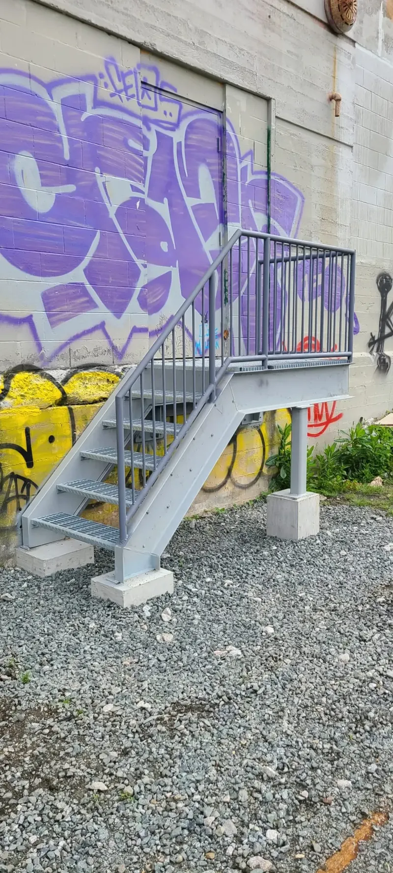 Short four-step galvanized platform stair with bar grating treads, pipe-rail guards, and concrete post bases, a back-entry warehouse replacement at a graffitied light-industrial building