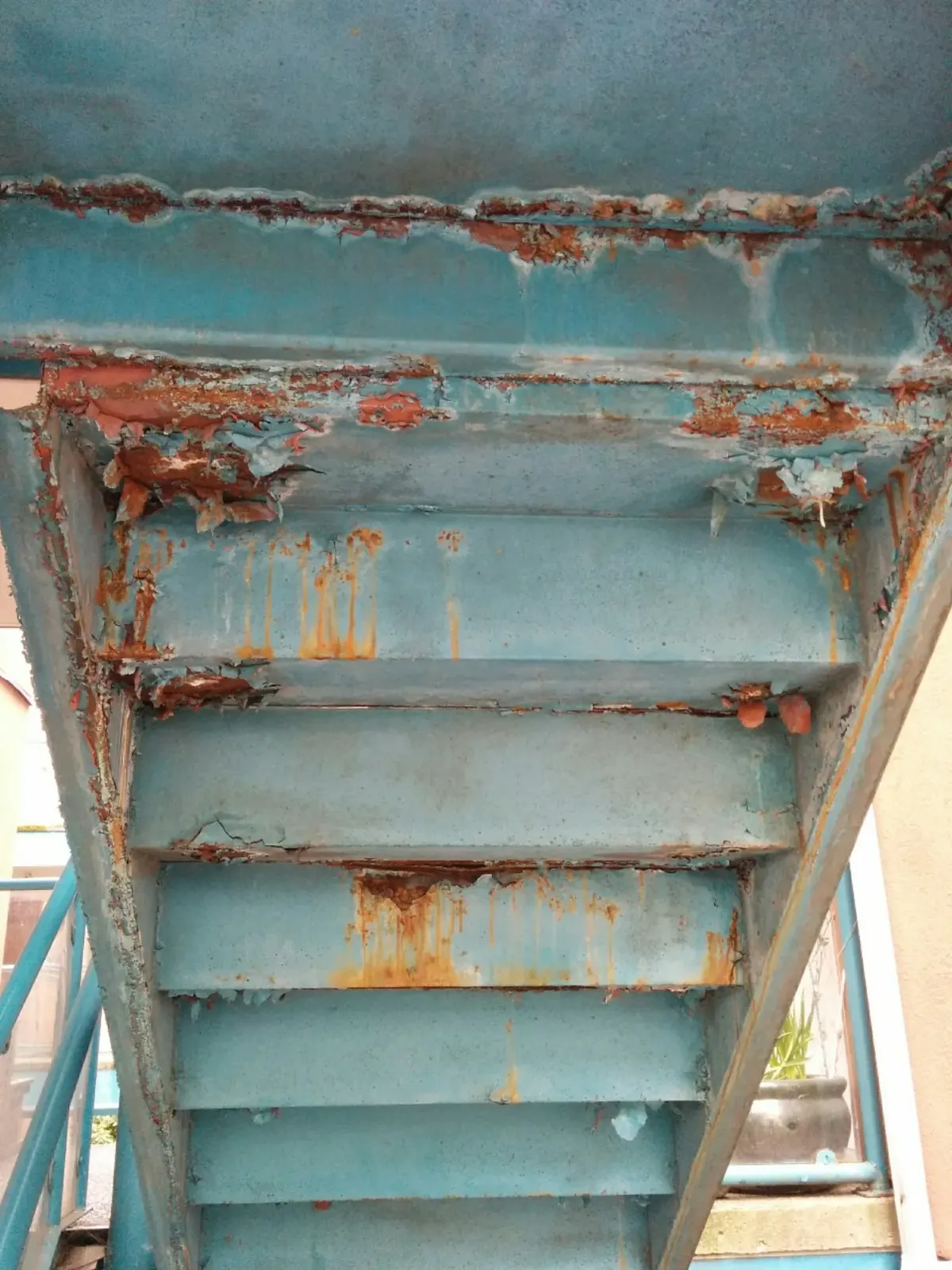 Underside of a corroded blue-painted exterior stair showing severe rust through the stringer-to-tread welds, paint failure, and chloride staining from years of ice melt