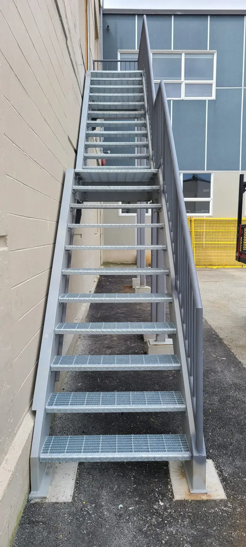Tall exterior steel access stair with galvanized bar grating treads against a Vancouver warehouse block wall, showing the open-drain detail that keeps water off the steel